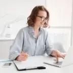 Smiling professional working at a laptop in a bright office, analyzing financial data with documents and a smartphone on the desk.