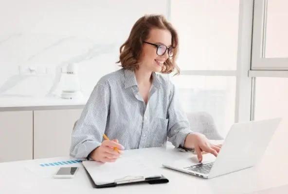 Smiling professional working at a laptop in a bright office, analyzing financial data with documents and a smartphone on the desk.