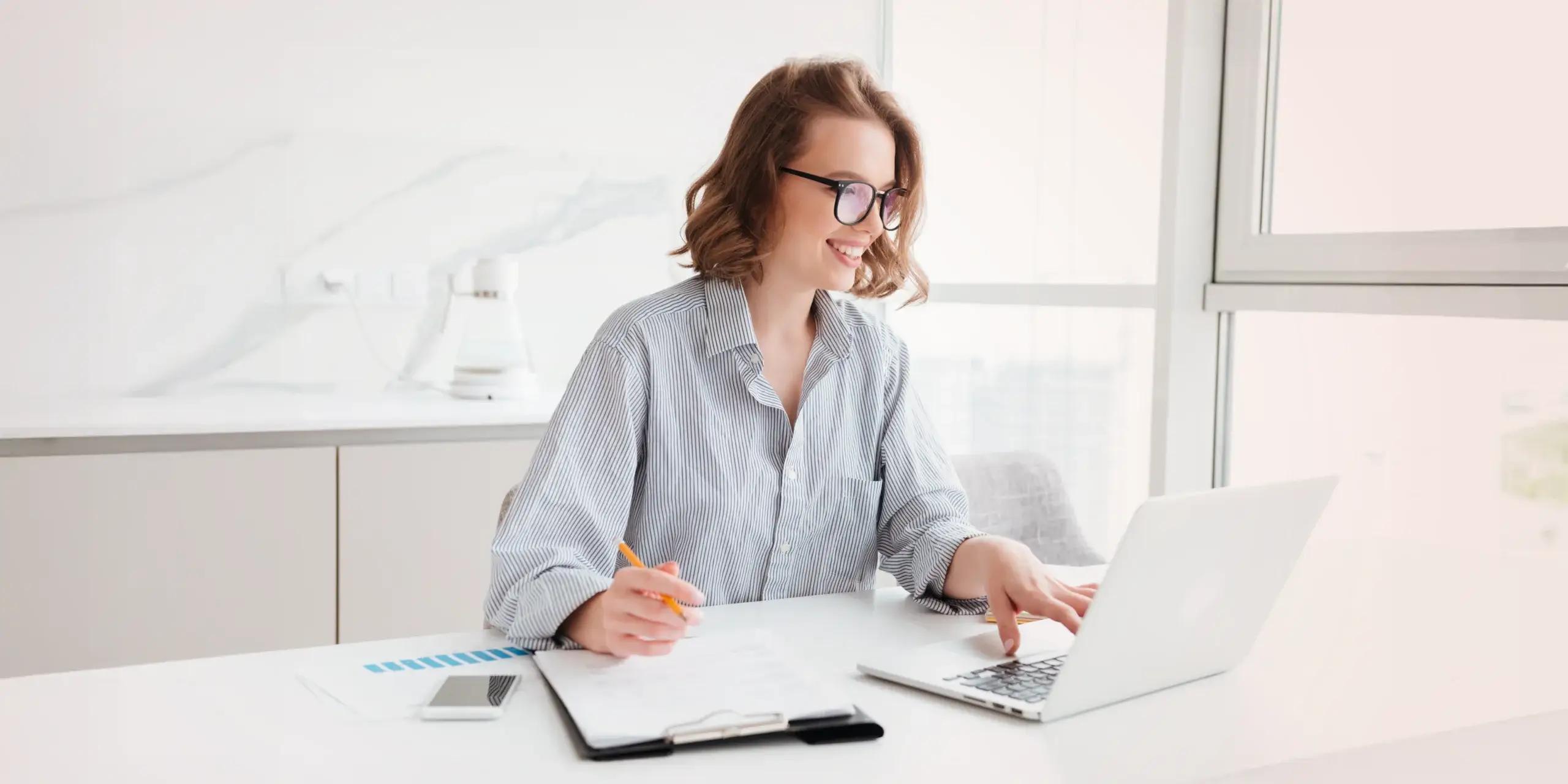 Smiling professional working at a laptop in a bright office, analyzing financial data with documents and a smartphone on the desk.