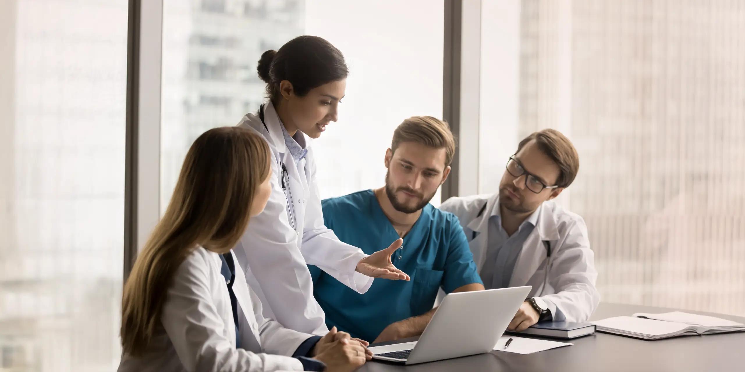 Medical professionals gathered in a meeting discussing patient care and practice strategies around a laptop in a hospital office.