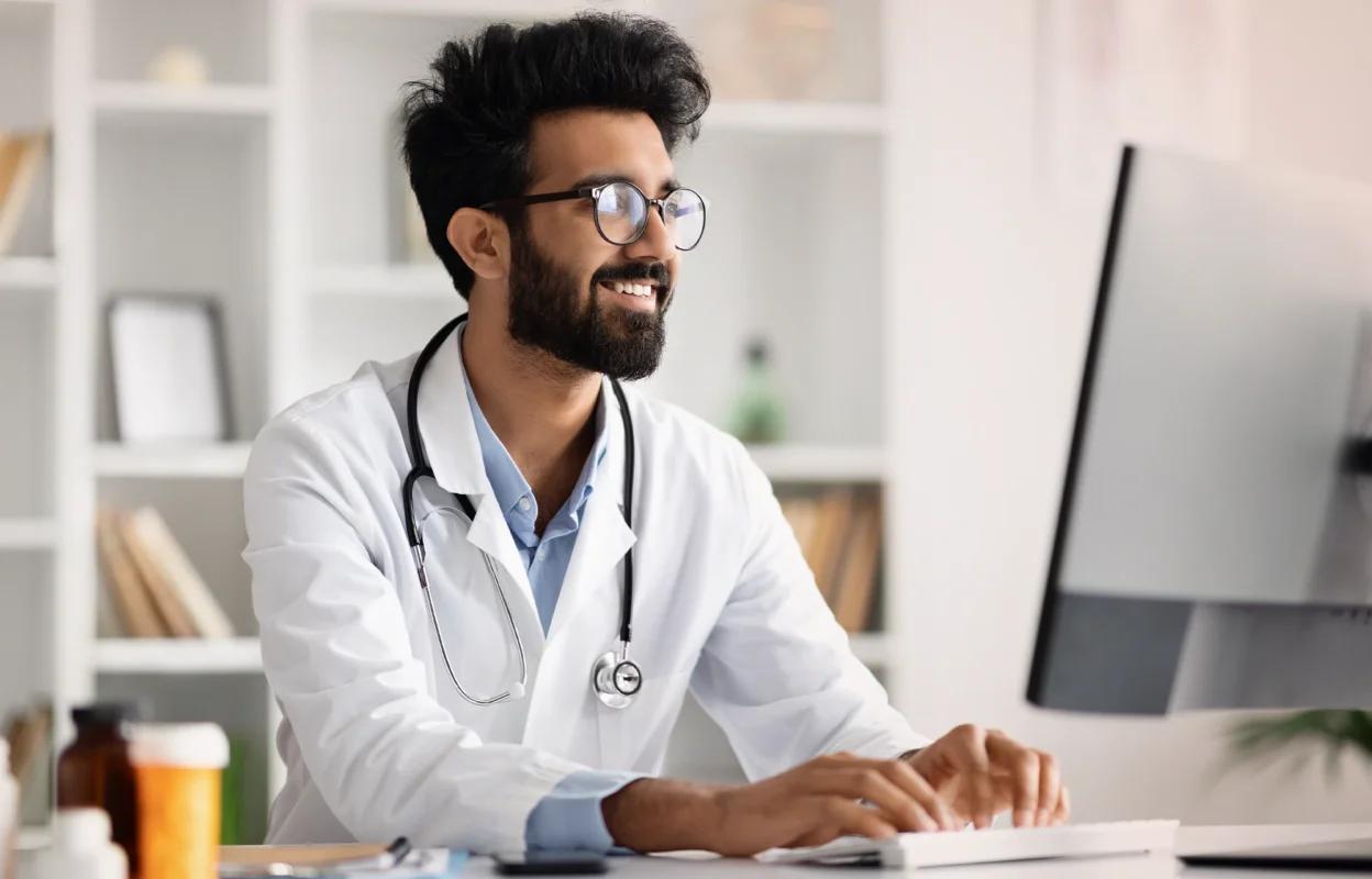 Smiling male doctor with a beard and glasses, wearing a white coat and stethoscope, sits at a desk typing on a computer in a well-lit office.