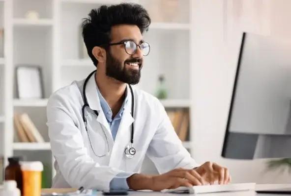 Smiling male doctor with a beard and glasses, wearing a white coat and stethoscope, sits at a desk typing on a computer in a well-lit office.