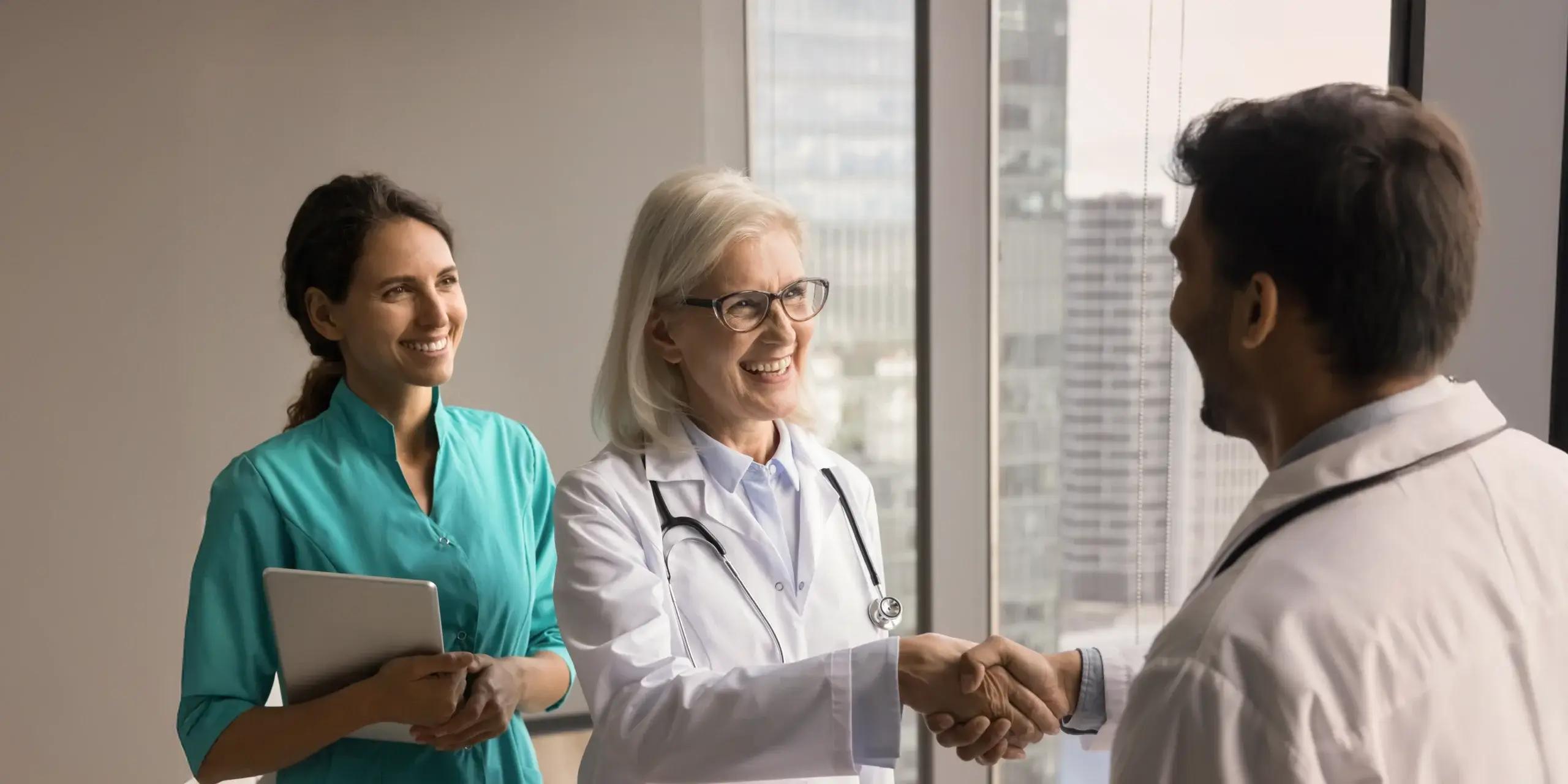 Smiling senior doctor shaking hands with a colleague while another medical professional looks on in a hospital office.