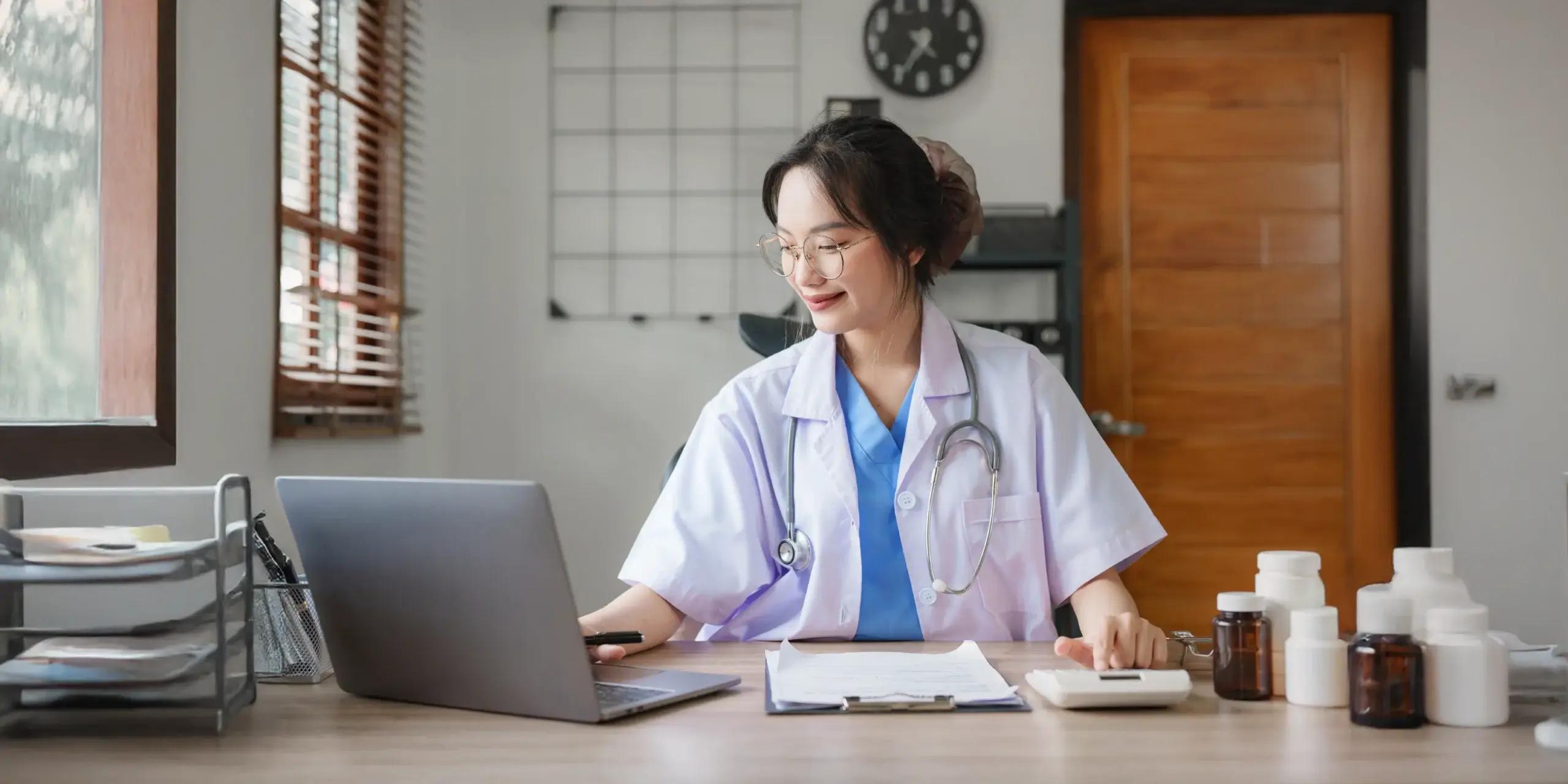 Smiling female doctor in an office reviewing medical notes on her laptop with medication bottles and paperwork on the desk.