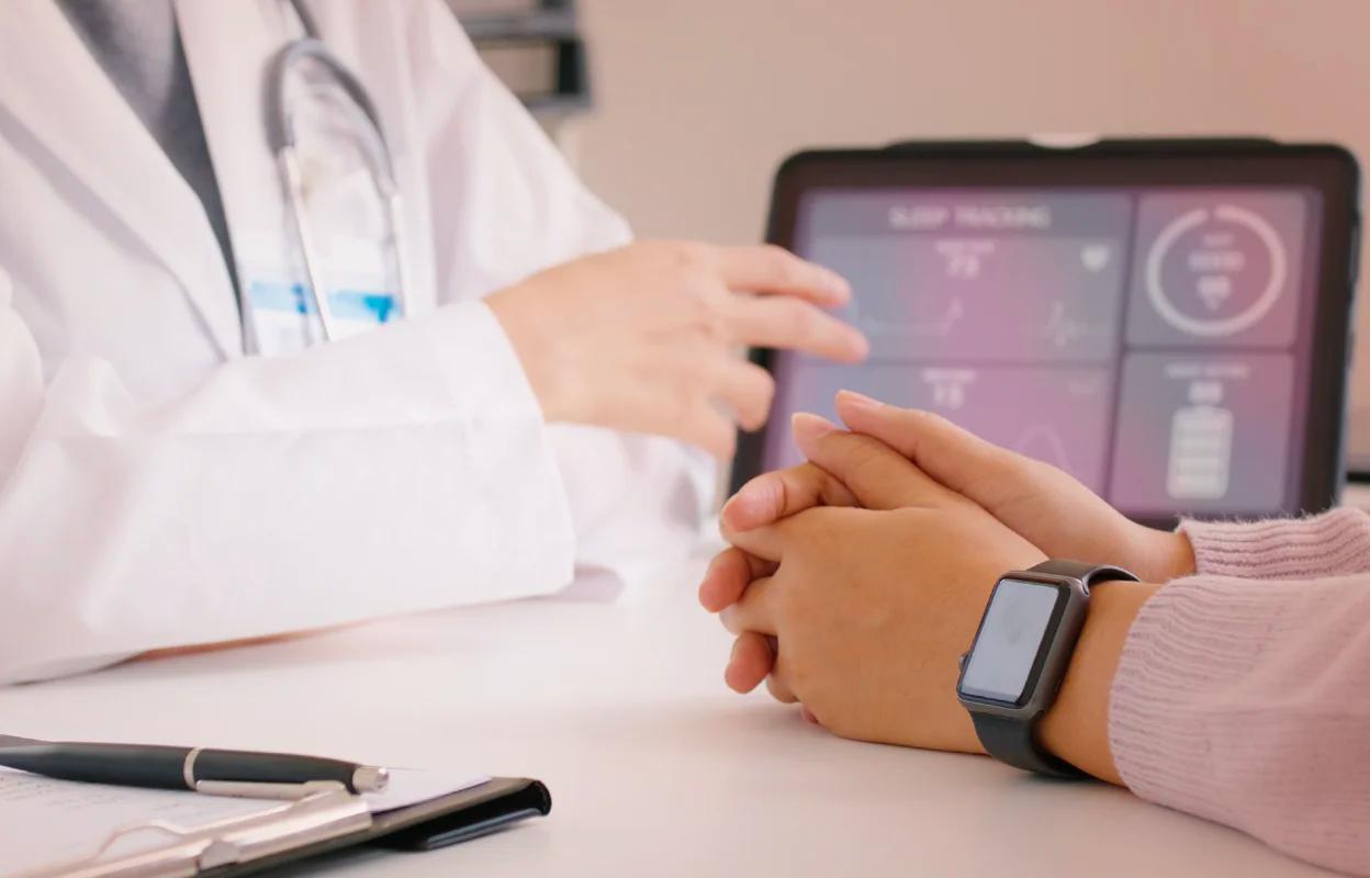 A doctor in a white coat is speaking with a patient seated across a desk, whose hands are clasped and wearing a smartwatch. A tablet on the desk displays health metrics like sleep tracking and heart rate in a dashboard format.