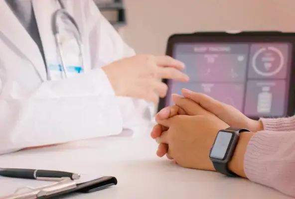 A doctor in a white coat is speaking with a patient seated across a desk, whose hands are clasped and wearing a smartwatch. A tablet on the desk displays health metrics like sleep tracking and heart rate in a dashboard format.