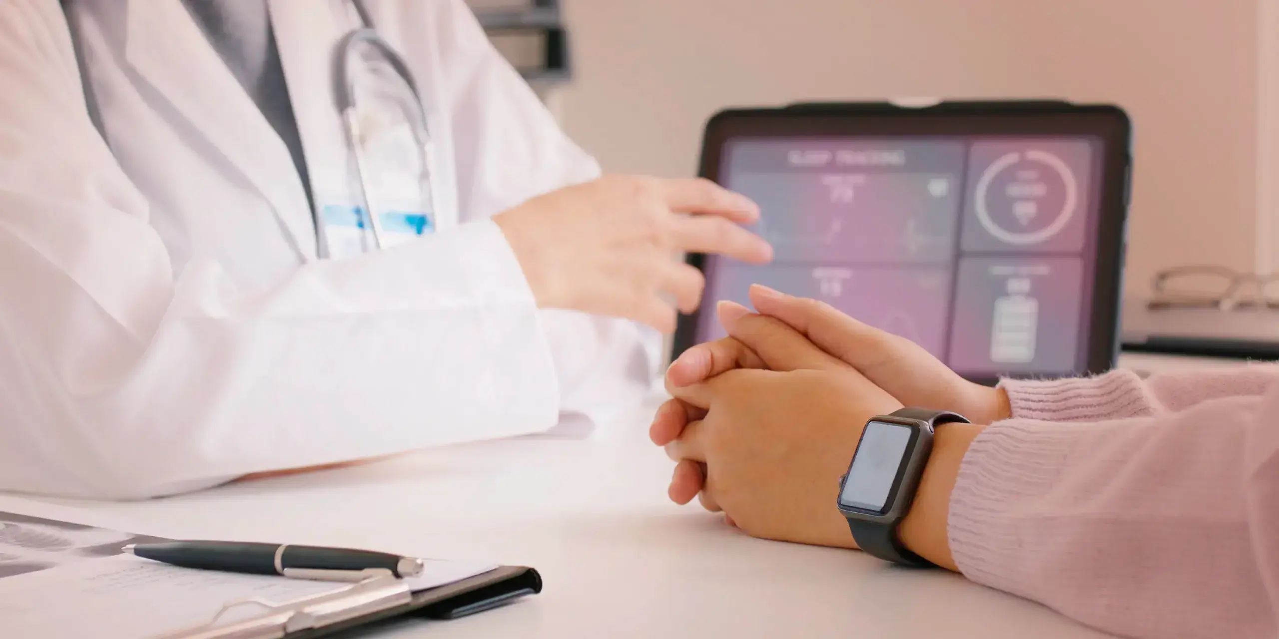 A doctor in a white coat is speaking with a patient seated across a desk, whose hands are clasped and wearing a smartwatch. A tablet on the desk displays health metrics like sleep tracking and heart rate in a dashboard format.
