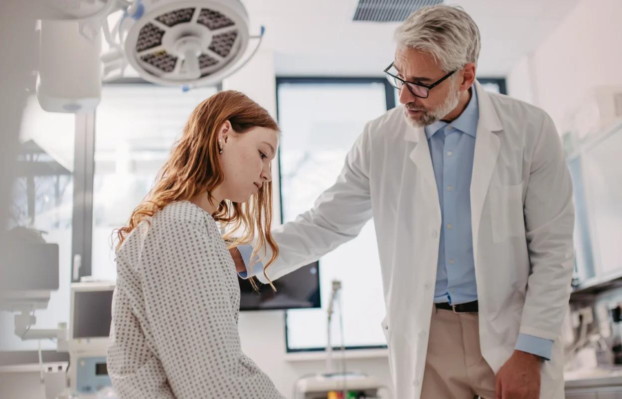 A middle-aged male doctor in a white coat gently consoles a young female patient in a hospital gown, seated on an exam table in a brightly lit medical room.