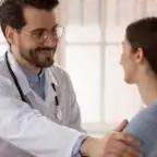 A male doctor in a white coat and stethoscope smiles warmly at a female patient while resting a reassuring hand on her shoulder during a consultation.