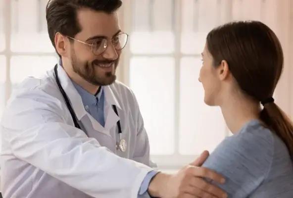 A male doctor in a white coat and stethoscope smiles warmly at a female patient while resting a reassuring hand on her shoulder during a consultation.