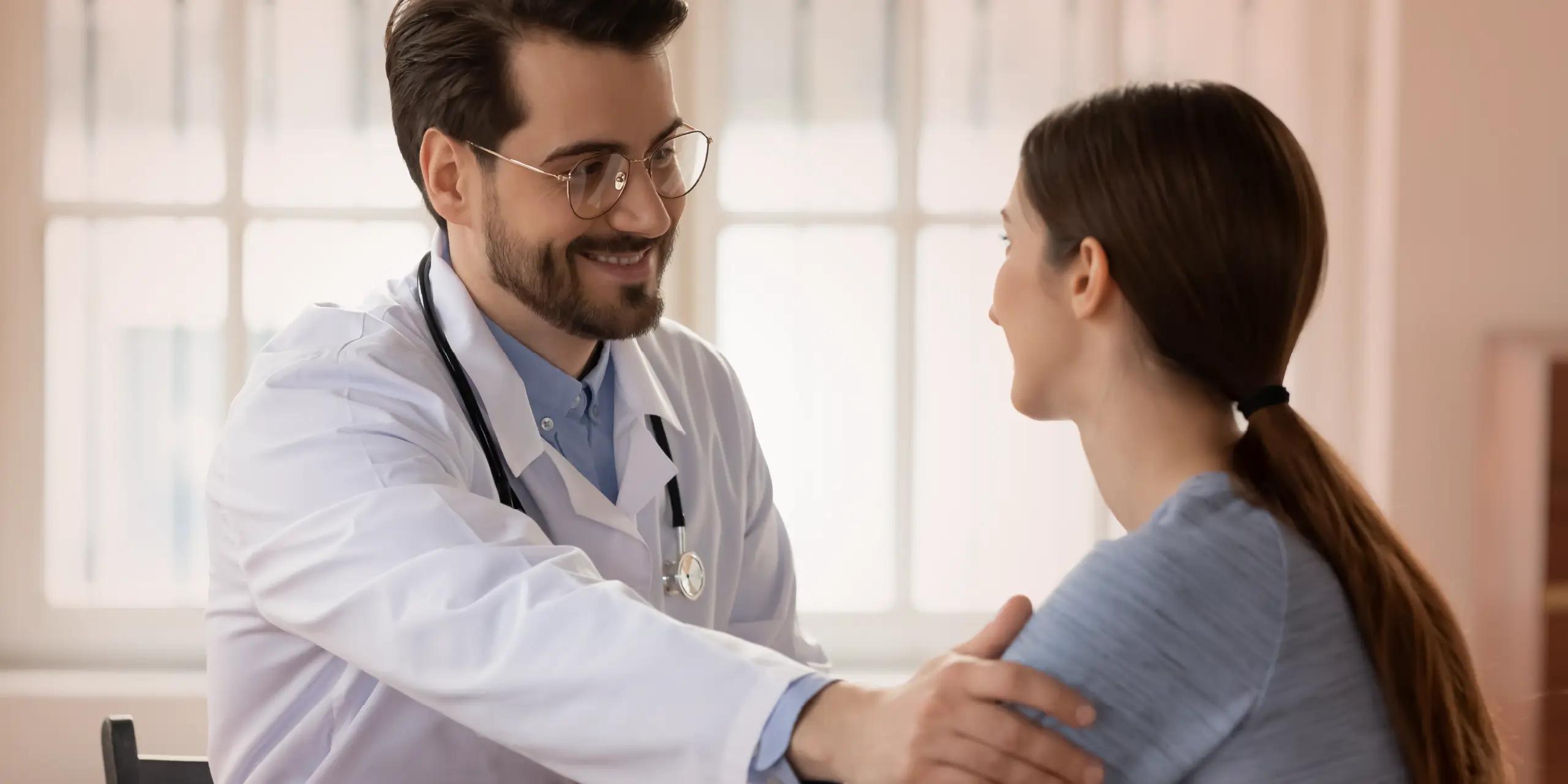 A male doctor in a white coat and stethoscope smiles warmly at a female patient while resting a reassuring hand on her shoulder during a consultation.