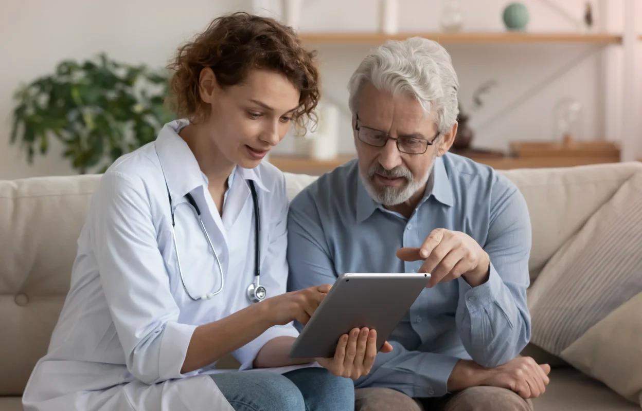 A female doctor and an older male patient sit on a couch, reviewing AI healthcare information on a tablet together.