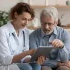A female doctor and an older male patient sit on a couch, reviewing information on a tablet together in a calm, home-like setting.
