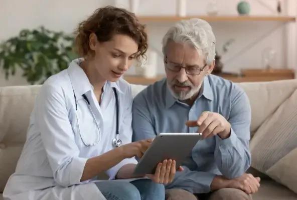A female doctor and an older male patient sit on a couch, reviewing information on a tablet together in a calm, home-like setting.