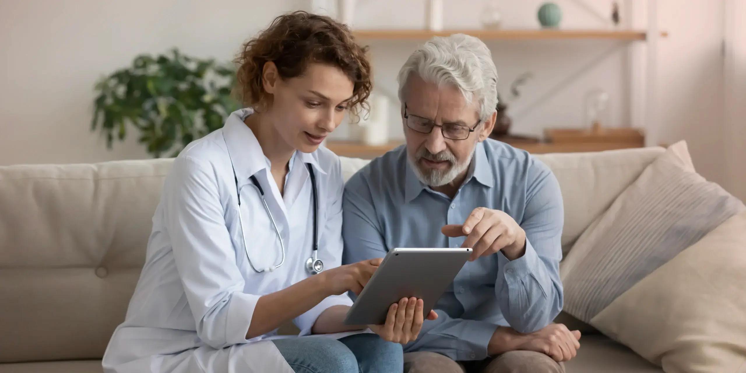 A female doctor and an older male patient sit on a couch, reviewing information on a tablet together in a calm, home-like setting.