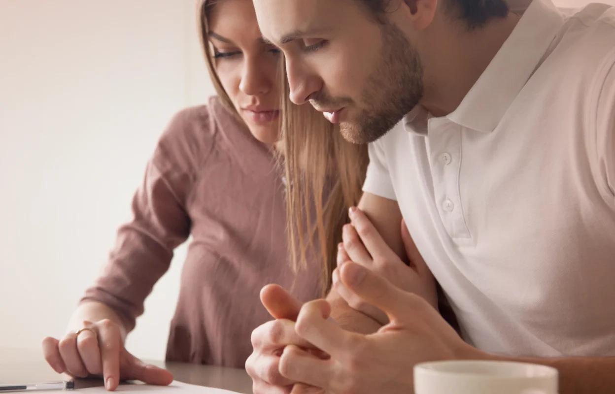 The image shows a couple sitting closely together at a table, looking concerned as they review paperwork, with a coffee cup and pen in front of them.