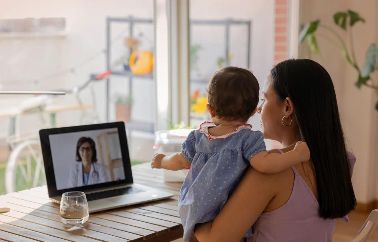 Image shows a woman on vacation holding a baby while on a video call with a doctor on her laptop at a wooden table.