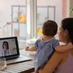 Image shows a woman on vacation holding a baby while on a video call with a doctor on her laptop at a wooden table.