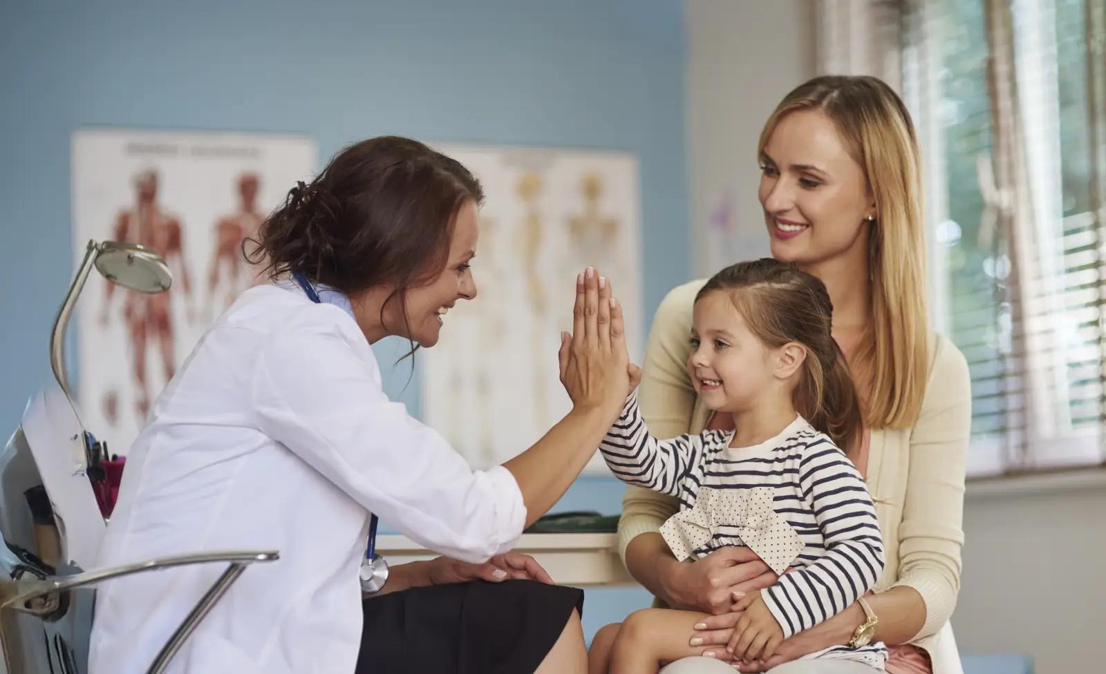 A female doctor smiles and gives a high five to a young girl sitting on her mother’s lap in a medical office.