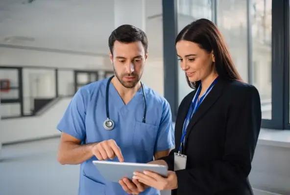 A clinician in scrubs and an administrator in professional attire look at a tablet together inside a bright medical facility.