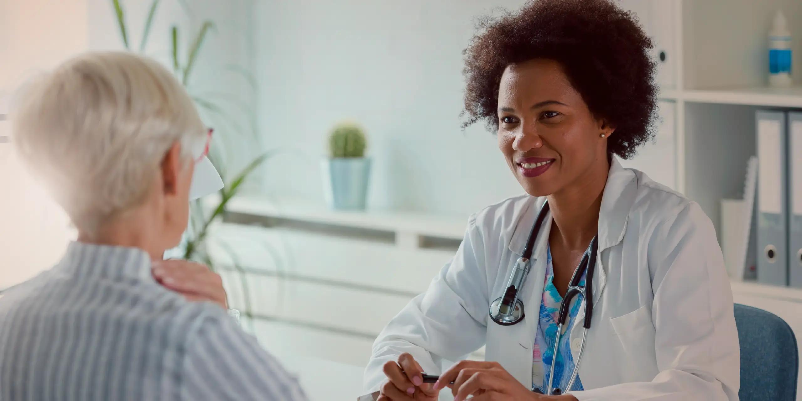 Smiling female doctor with a stethoscope talks to an older woman patient in a medical office.