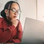 Woman wearing glasses sits at a desk using a laptop, focused on reviewing healthcare or medical billing information in a bright, modern workspace.