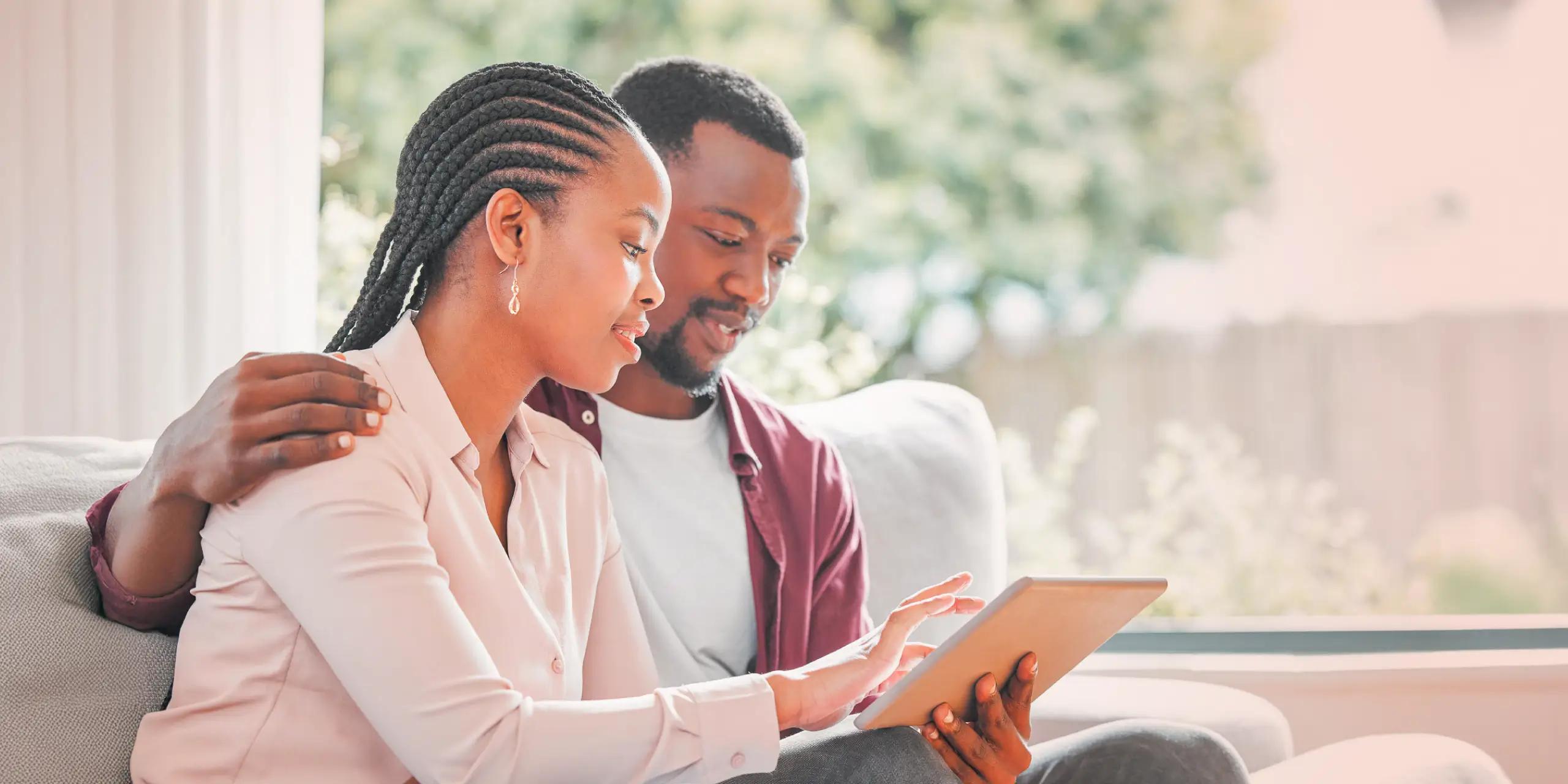 A couple sitting together on a couch, reviewing healthcare information on a tablet in a bright living room.