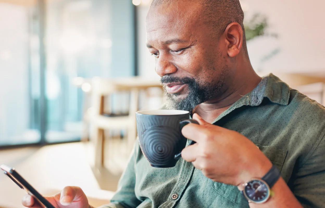 Middle-aged man sitting indoors, holding a coffee mug and looking at his smartphone in a bright, modern setting.