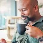 Middle-aged man sitting indoors, holding a coffee mug and looking at his smartphone in a bright, modern setting.