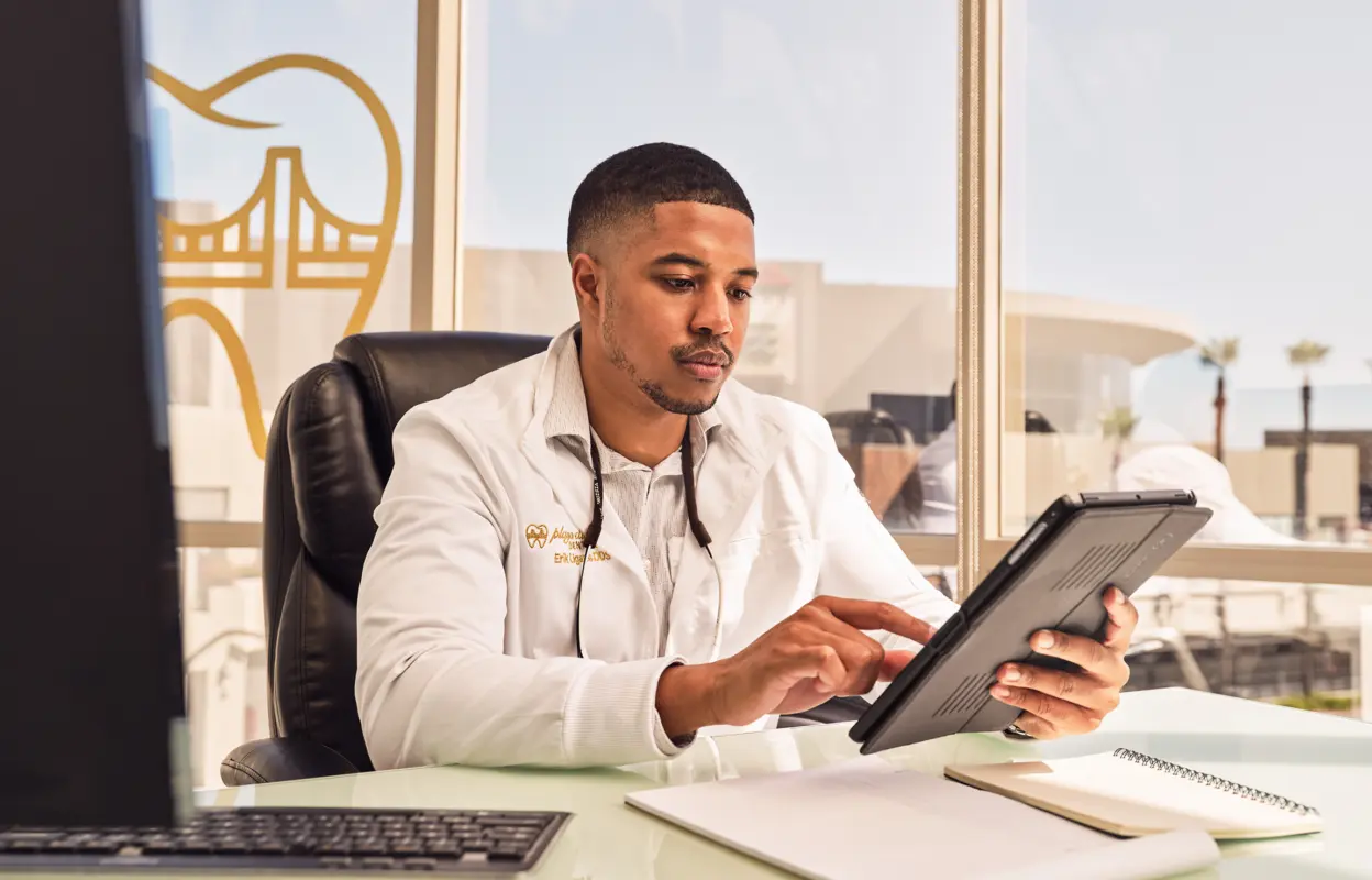 Male dentist wearing a white coat working on a tablet at his desk with dental equipment and office window in the background.