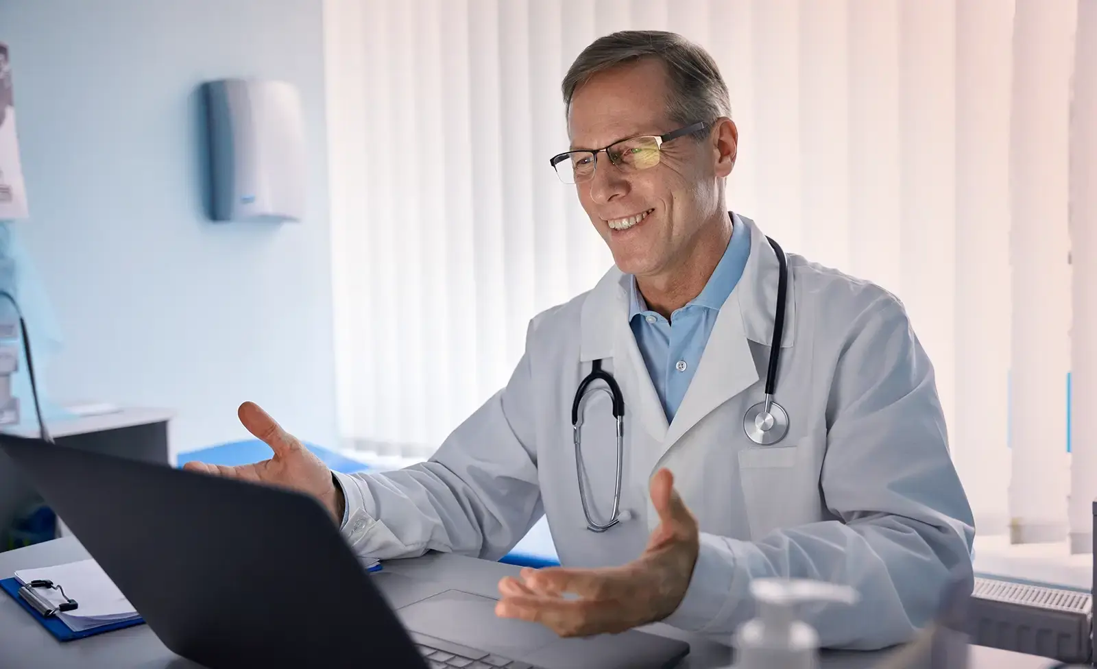Male doctor wearing a white coat and stethoscope gestures while smiling during a video call on a laptop.
