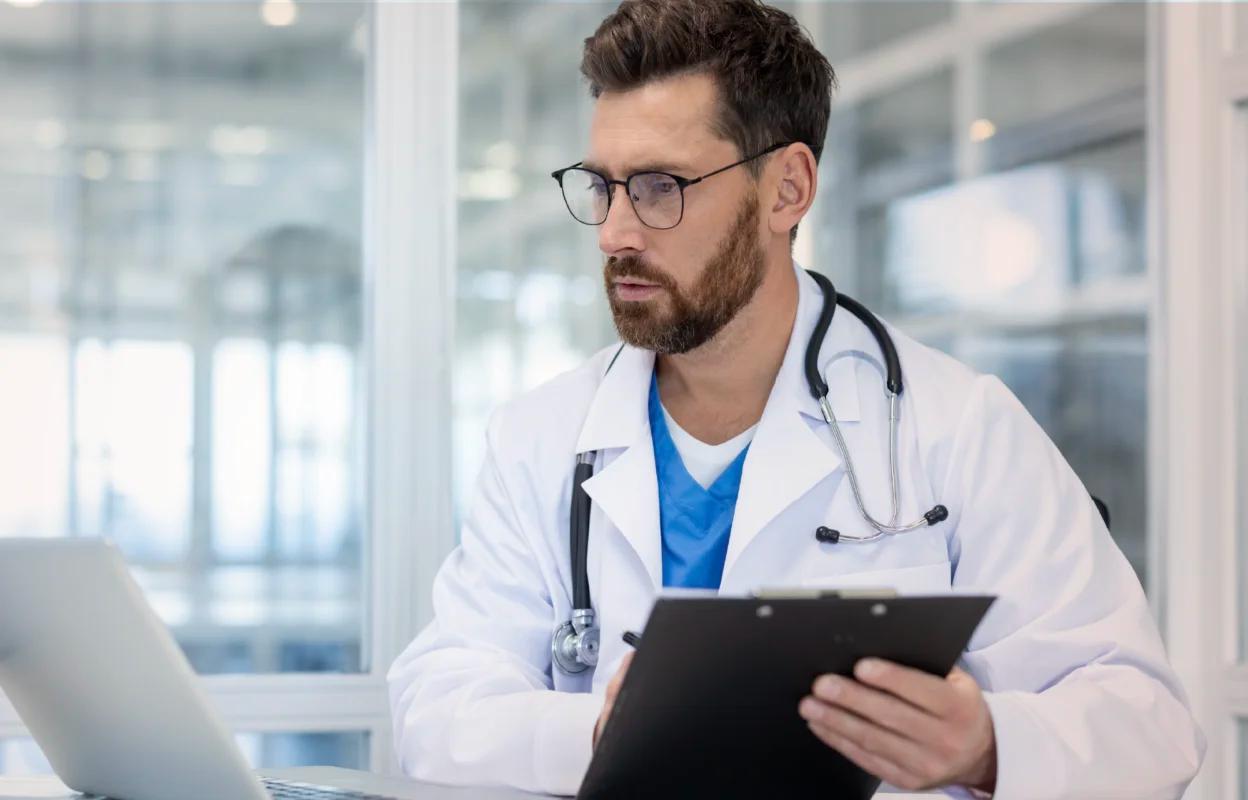 Male doctor with glasses and a stethoscope around his neck sitting in a bright office, holding a clipboard and looking at a laptop.