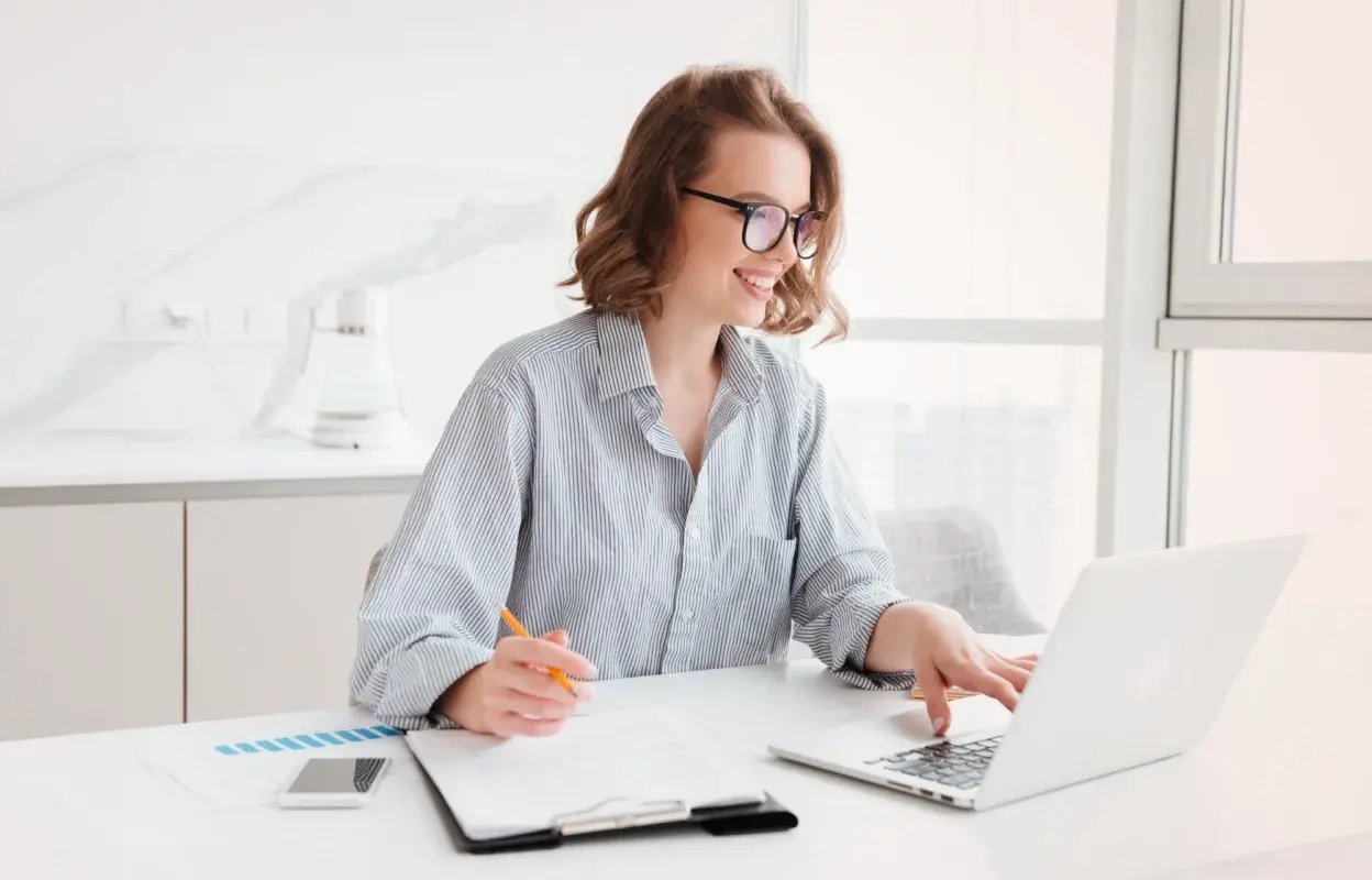 Smiling professional working at a laptop in a bright office, analyzing financial data with documents and a smartphone on the desk.