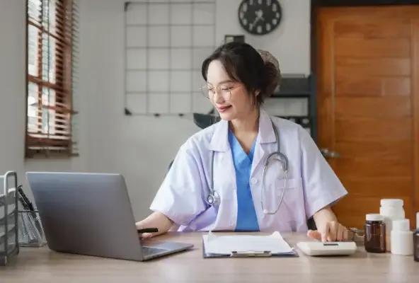 Smiling female doctor in an office reviewing medical notes on her laptop with medication bottles and paperwork on the desk.