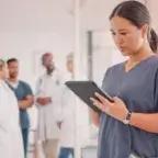 Thumbnail image from Tebra featuring a nurse in scrubs holding a tablet, with a blurred background of medical staff, highlighting themes of reflection and stress in the nursing profession.
