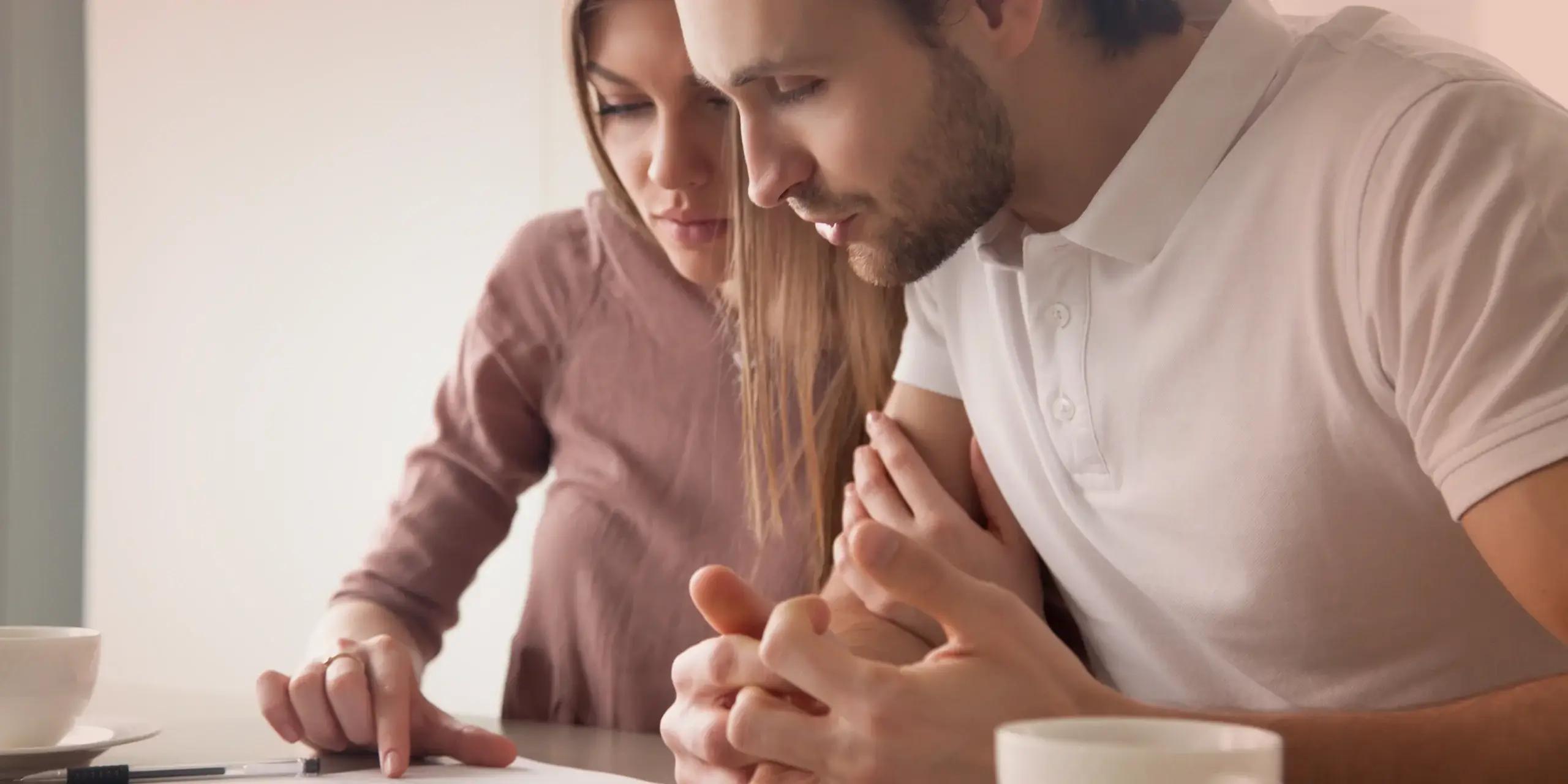 The image shows a couple sitting closely together at a table, looking concerned as they review paperwork, with a coffee cup and pen in front of them.