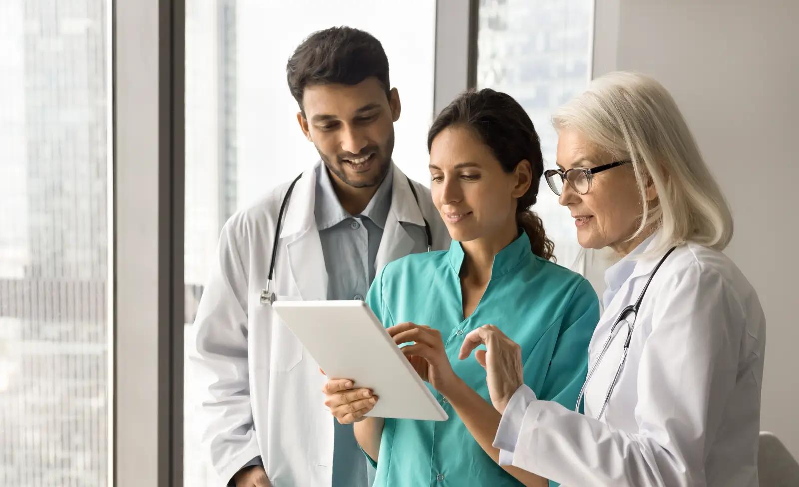 Three healthcare professionals stand together reviewing information on a tablet near a large window, smiling as they collaborate.