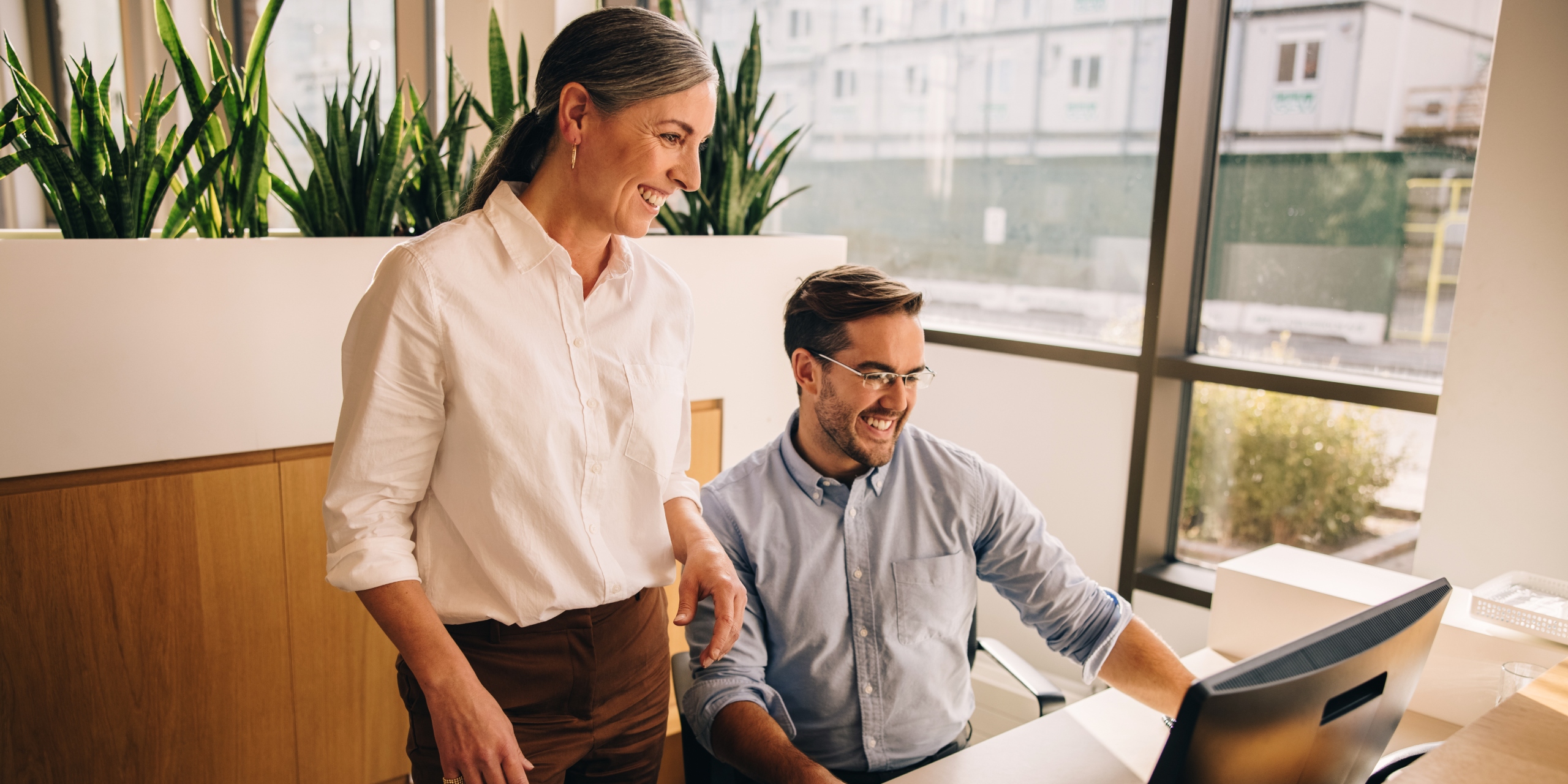 Two smiling colleagues in an office setting looking at the same computer.